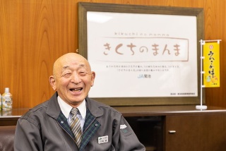 安心品質をうたうJA菊池のブランド「きくちのまんま」ロゴをバックに笑う三角氏。阿蘇山の裾野で培われた豊かな自然が市街地の人々の生活を支えている(写真右はJA菊池 本所外観)
