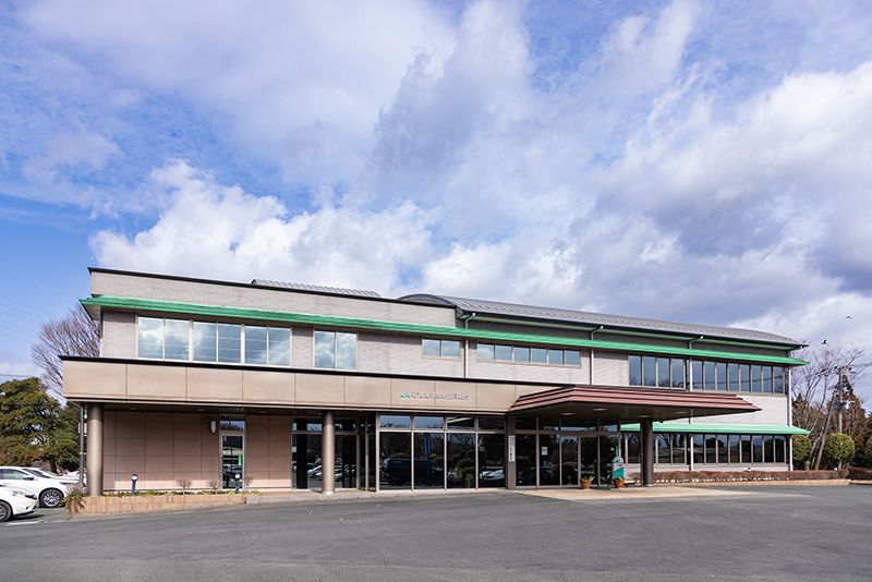 安心品質をうたうJA菊池のブランド「きくちのまんま」ロゴをバックに笑う三角氏。阿蘇山の裾野で培われた豊かな自然が市街地の人々の生活を支えている（写真右はJA菊池 本所外観）