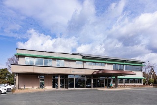 安心品質をうたうJA菊池のブランド「きくちのまんま」ロゴをバックに笑う三角氏。阿蘇山の裾野で培われた豊かな自然が市街地の人々の生活を支えている(写真右はJA菊池 本所外観)