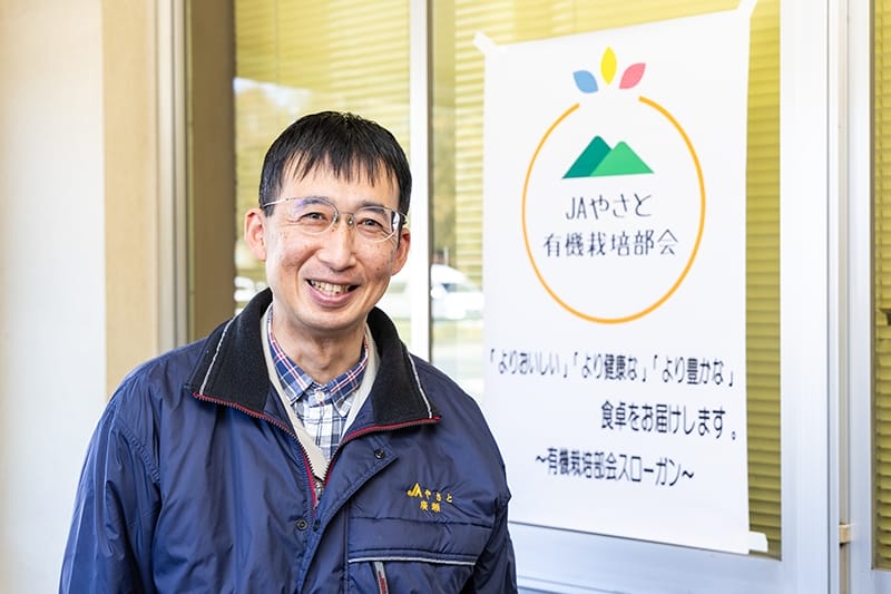 有機栽培部会のロゴとスローガンを背景に、笑顔の廣瀬氏。ロゴは部会に所属する生産者からデザインを募集したという