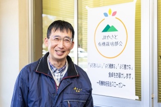 有機栽培部会のロゴとスローガンを背景に、笑顔の廣瀬氏。ロゴは部会に所属する生産者からデザインを募集したという