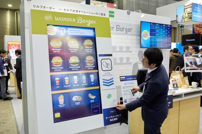 ブースでは、セルフ決済システムの体験デモも実施。会員情報を記録したカードをかざすと、その人に合ったメニューが画面に表示され、決済を行えるものだ。外国人向けには言語を切替えて、車椅子の人向けにはメニュー表示を画面下の方へ移動させて表示。カードに入金しておけば、キャッシュレスでセルフ決済が可能で、混雑を緩和できる。